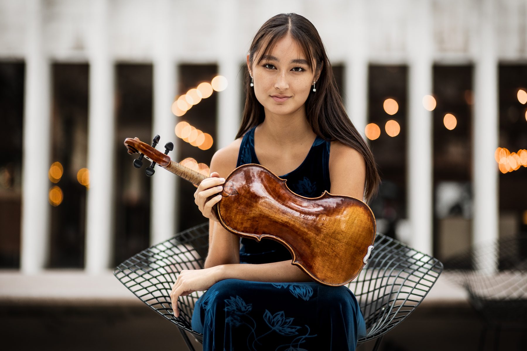 Maya Kilburn in a blue dress, seated with her violin
